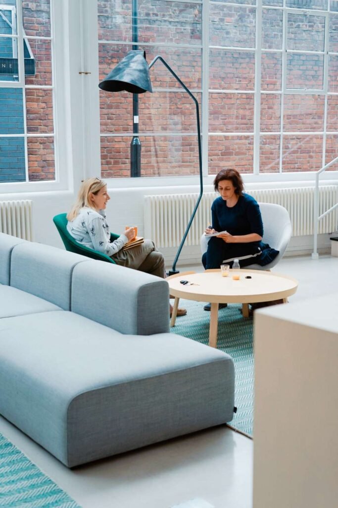 Two women sit in a bright, modern lounge by large windows, chatting over coffee at a round wooden table.