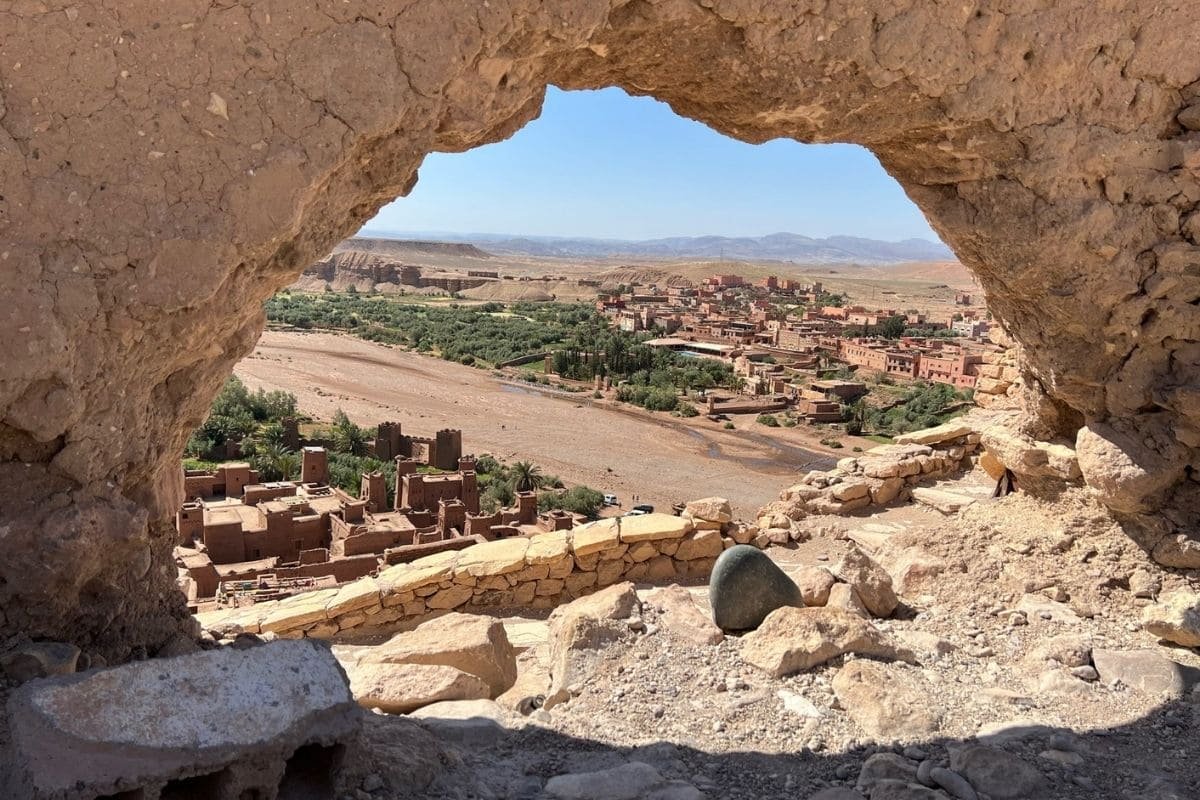View of a desert town framed by a rough stone arch in a rocky foreground, with mud-brick buildings and a river valley beyond.