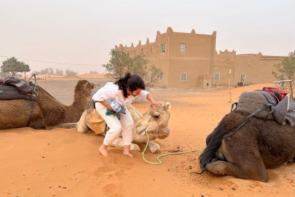 Woman in white clothing pets a camel in a desert camp, with a mud-brick building in the background and caravans nearby.