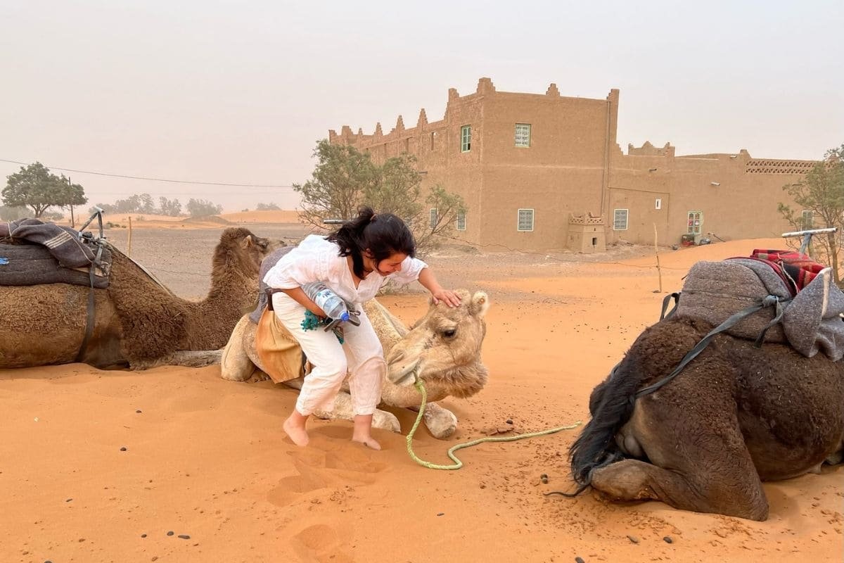 Woman in white clothing pets a camel in a desert camp, with a mud-brick building in the background and caravans nearby.