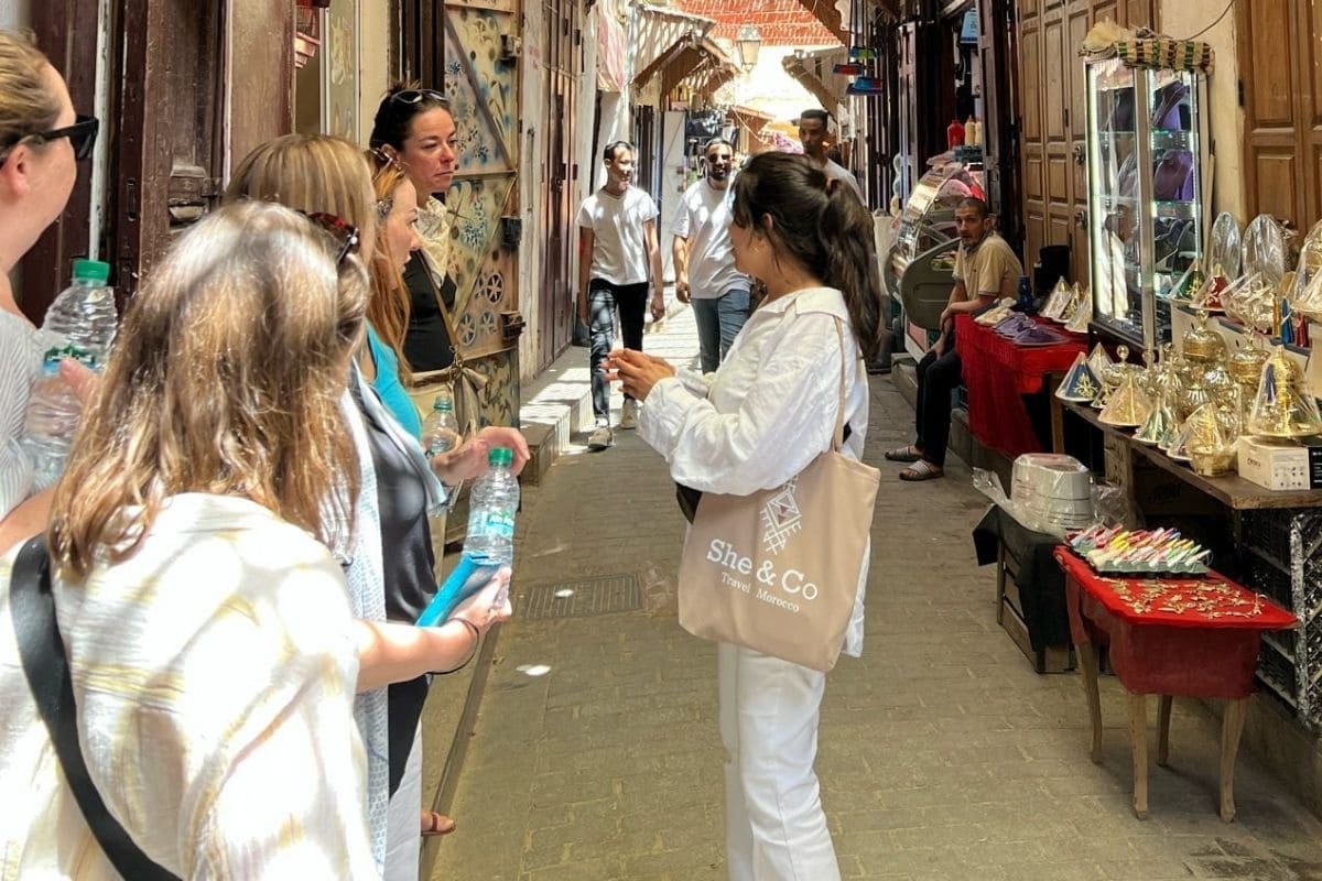 Group of tourists in a narrow Moroccan market street listening to a guide, souvenir stalls on the right side.