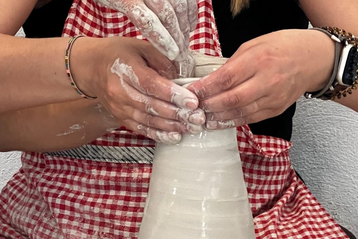 Two hands shape a tall white clay vase on a pottery wheel, both hands and wrists covered in wet clay, wearing red plaid aprons nearby.