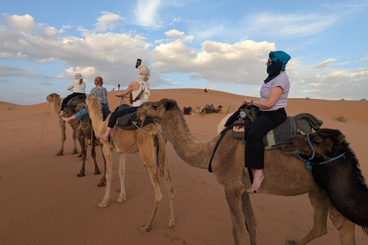 Group of tourists riding camels in a sandy desert under a blue sky with scattered clouds, wearing headscarves and light clothing.