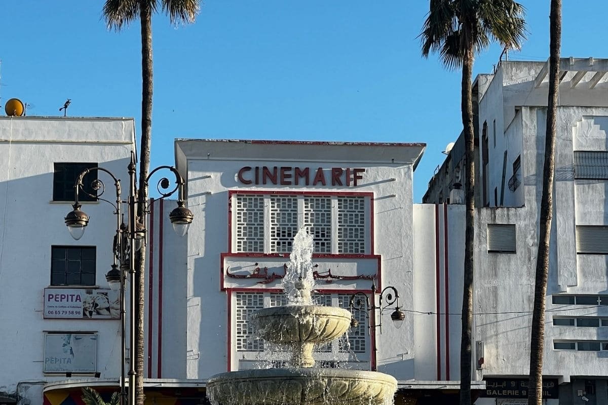 Art-deco cinema facade with a tiered fountain in front, flanked by palm trees and decorative street lamps on a sunny day.