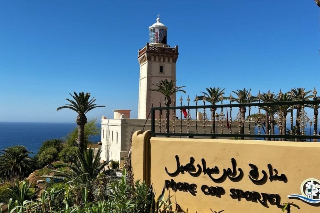 Coastal scene with a tall lighthouse, blue sea and sky, and palm trees beside a yellow sign with Arabic lettering in the foreground.