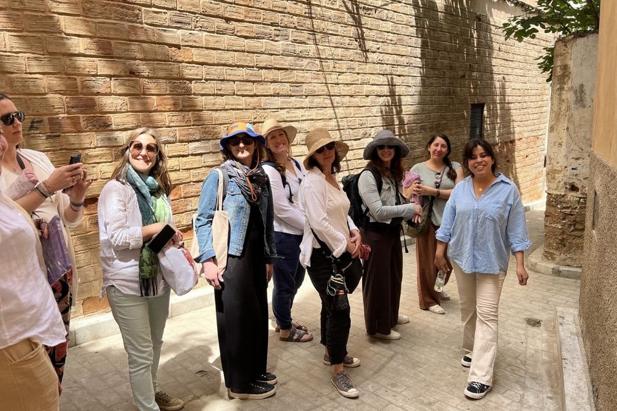 Group of women on a guided tour along a sunlit brick wall, wearing hats and casual clothing.