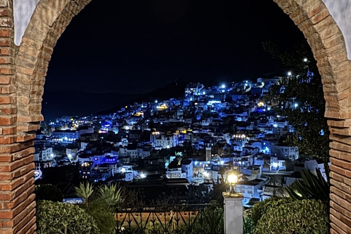 Night view of a hillside town lit with blue and warm lights, framed by a brick arch and foreground plants.