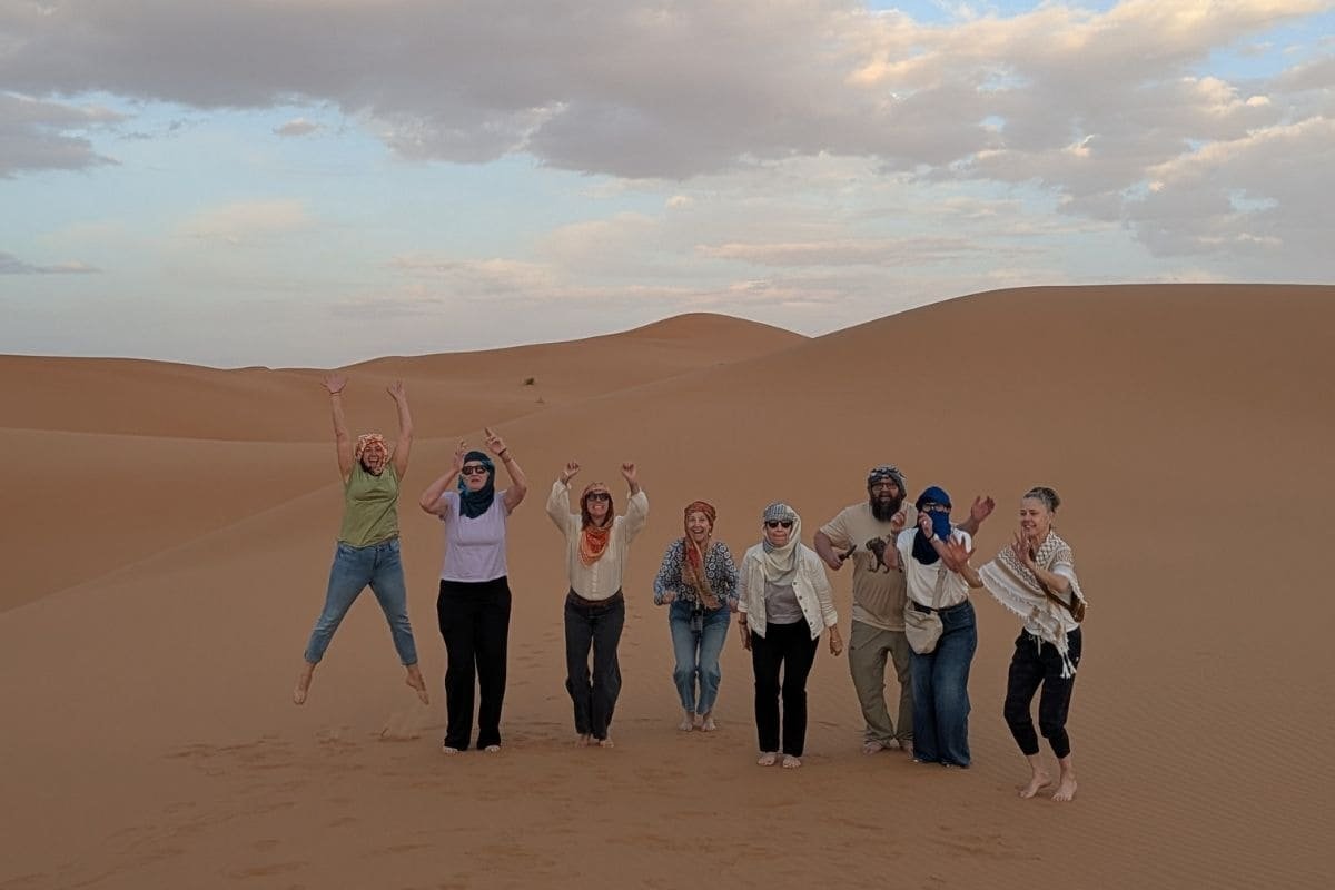 Group of friends standing on orange sand dunes, some jumping with arms raised, under a partly cloudy sky.