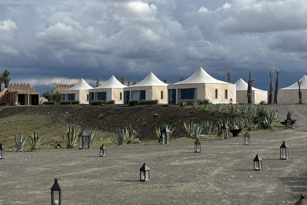 Row of white tents with pointed roofs on a sandy hill, desert plants in the foreground, and lanterns scattered across the ground under a cloudy sky.