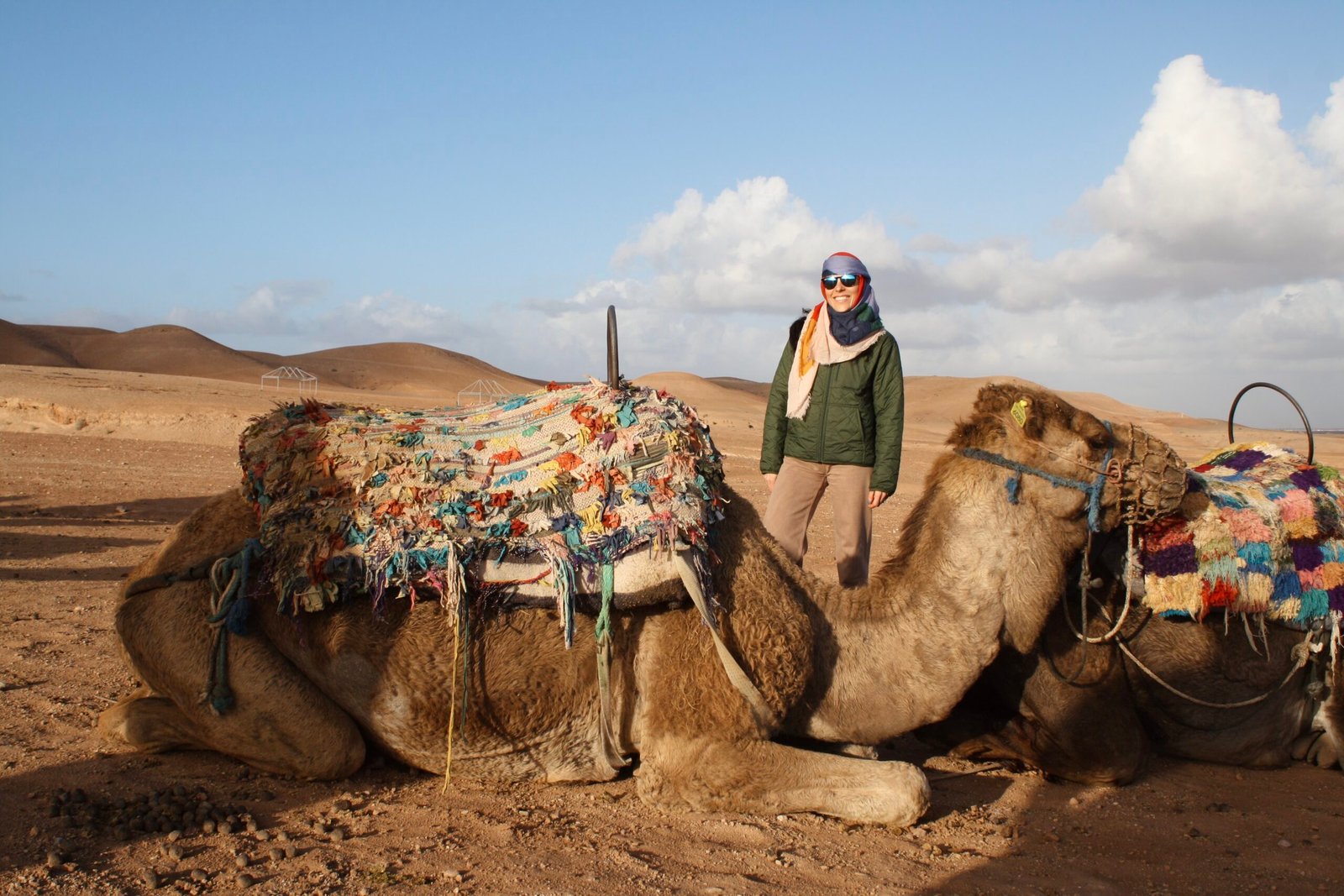 Person standing beside a camel in a desert, wearing a green jacket, scarf and sunglasses, with colorful woven saddles on the camel under a blue sky.