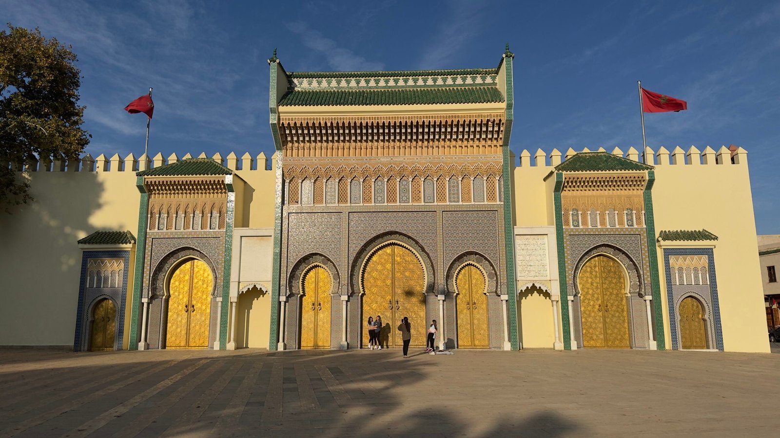 Moroccan-style entrance with gold doors, green tilework, and a yellow wall, flanked by red flagpoles; a few people stand at the doorway.