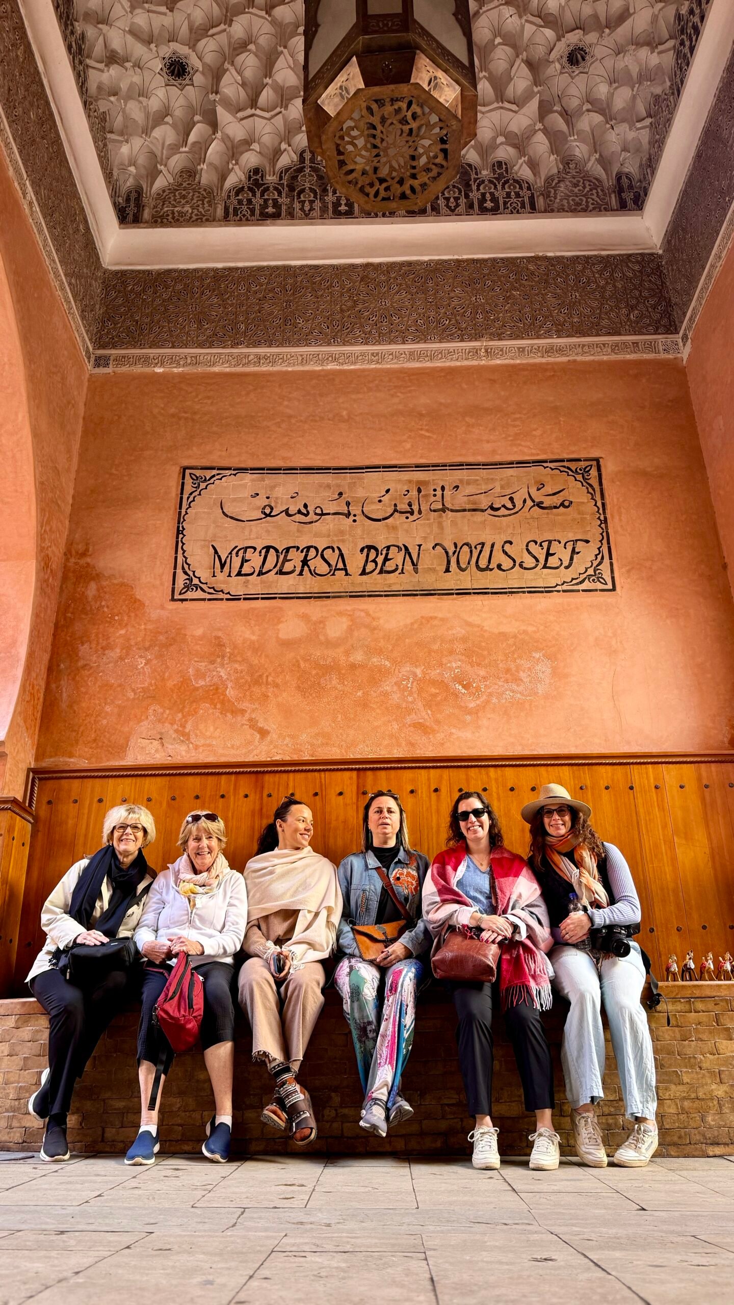 Seven women pose and sit on a wooden bench inside a warmly lit, ornately decorated hall with a sign that reads 'Medersa Ben Youssef'
