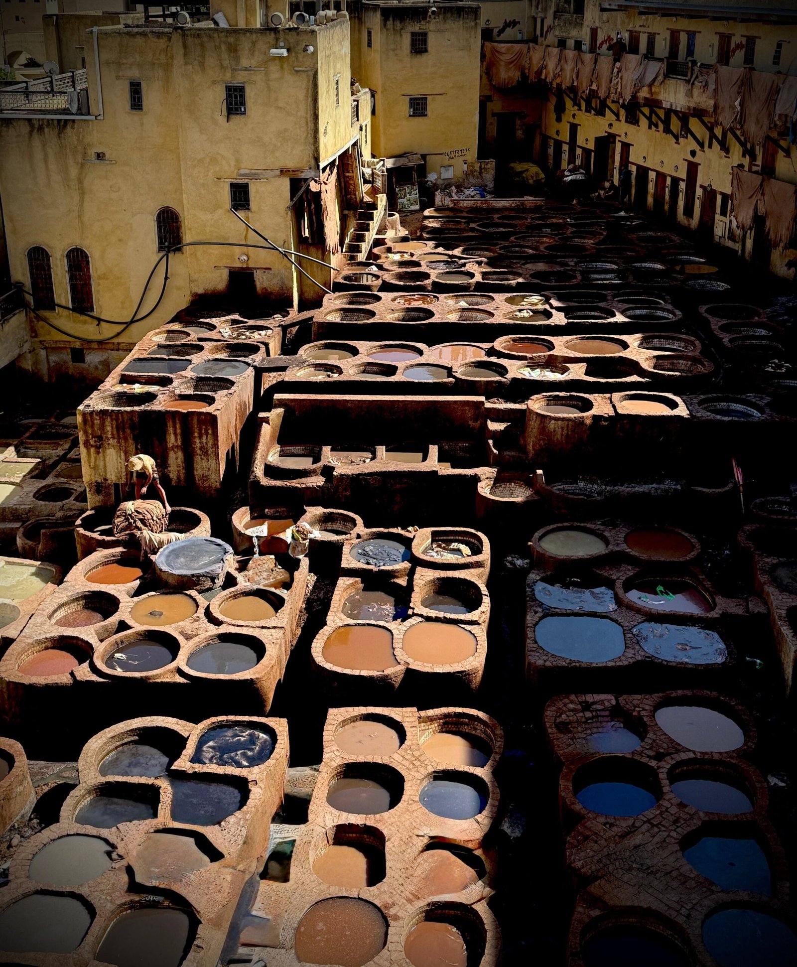 Open-air leather tannery with rows of circular dye-filled vats and yellow stucco buildings surrounding the pits