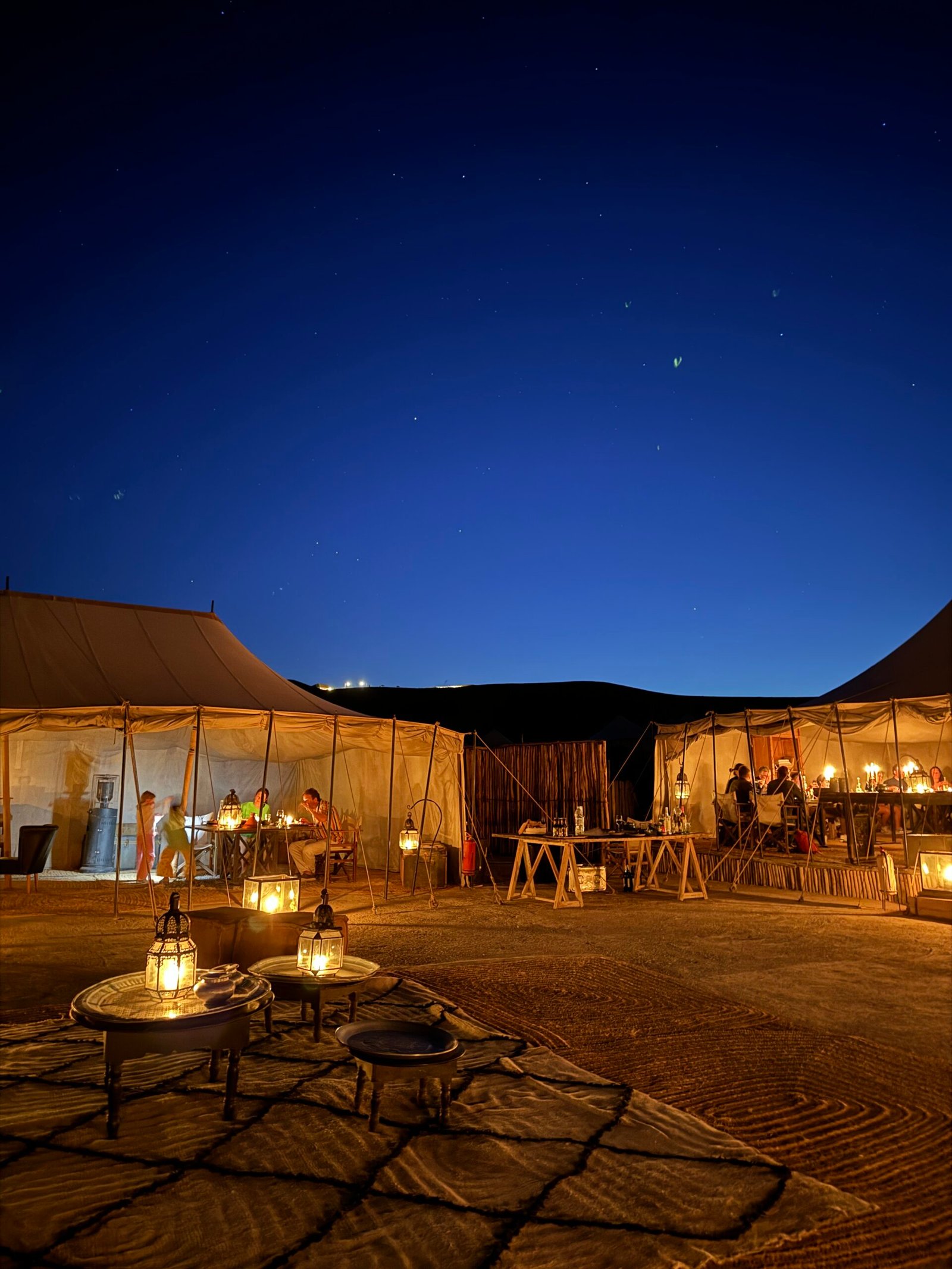Desert campsite at night with beige tents, lantern-lit tables, and people dining under a starry sky.